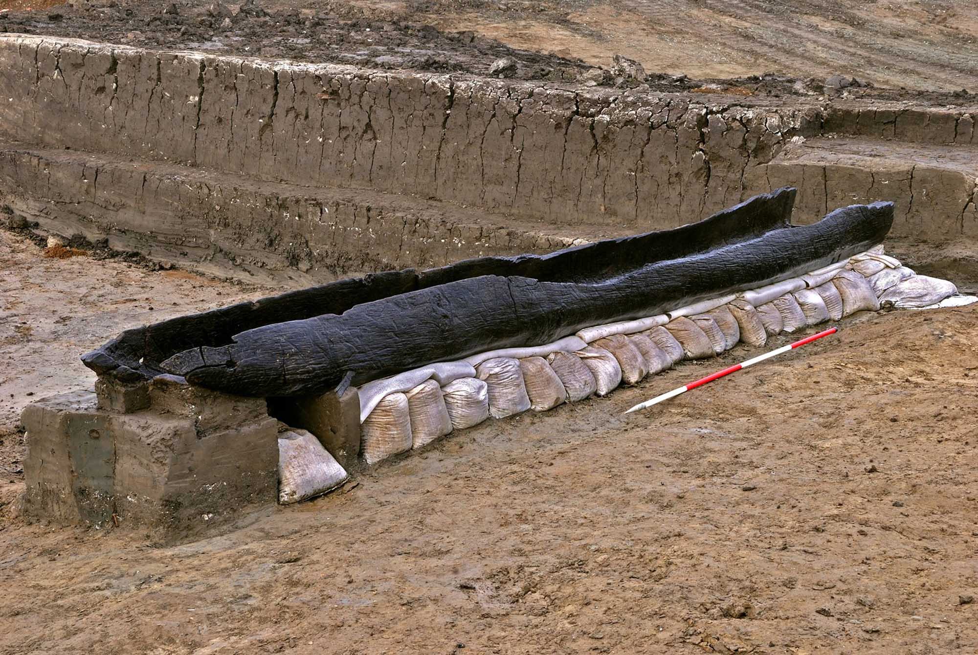 A hollowed, blackened tree log resting on a line of sand bags with a measuring stick alongside.