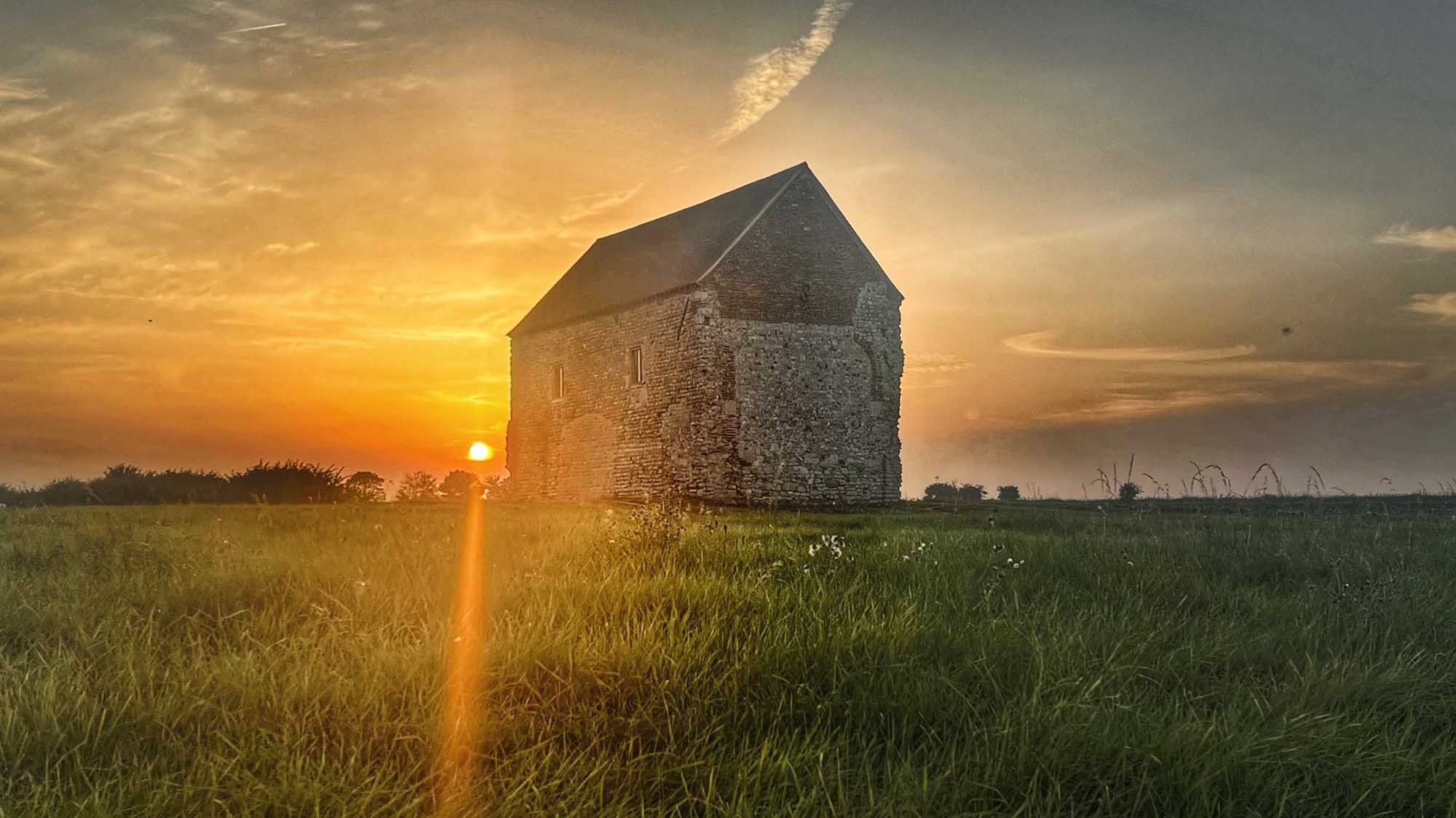 A stone monastery in the middle of a grassy field with the sun setting behind.