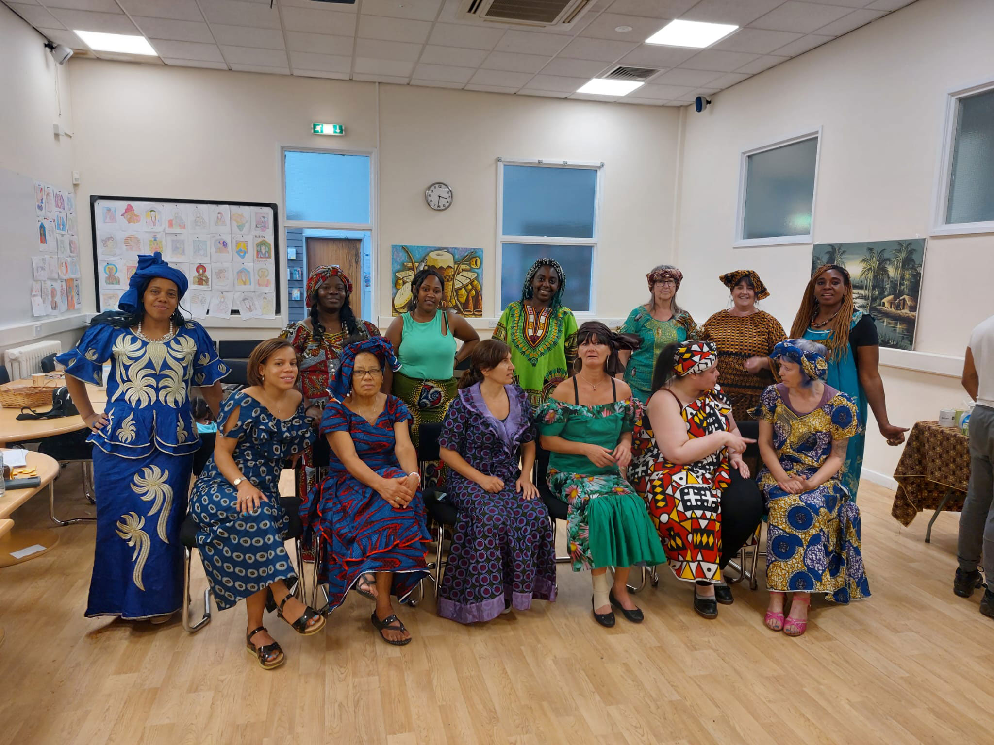 A group of women in two rows. The row at the back is standing while the row at the front is seated. They are wearing colourful bold print dresses posing for the photo. 
