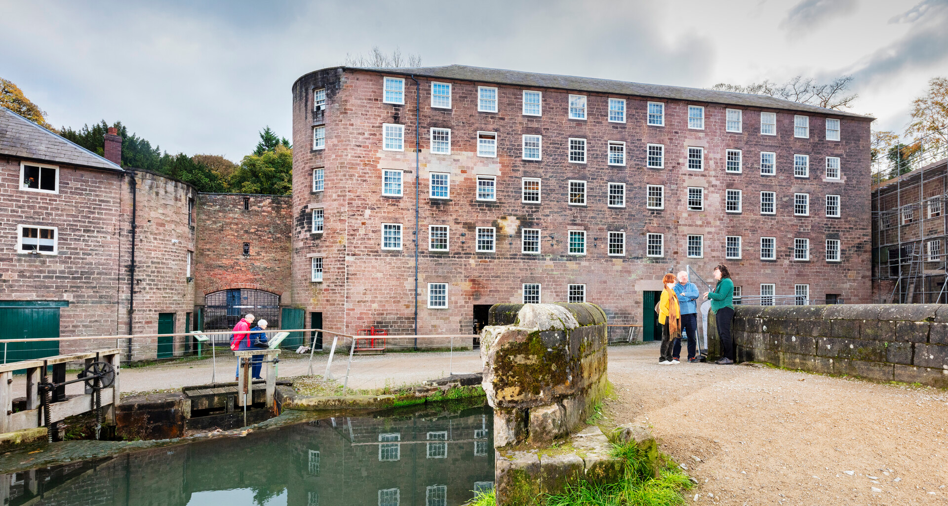 A historic stone mill building beside a calm waterway, with a small bridge in the foreground where 3 people are standing having a conversation. 
