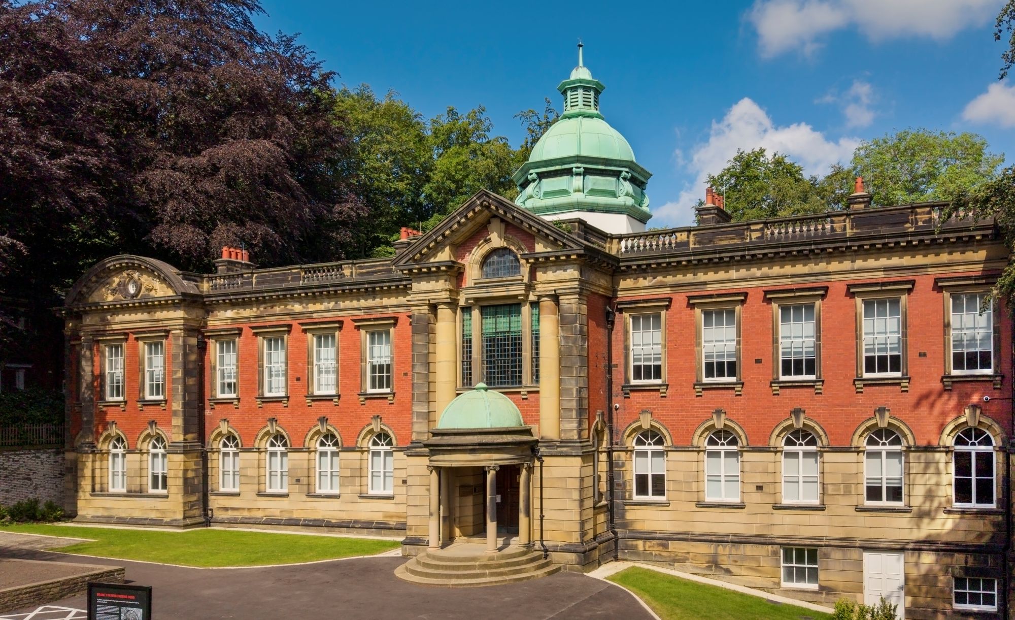 Front view of a large red-brick historic building with a central green-domed roof, arched windows and a grand entrance surrounded by trees under a blue sky.