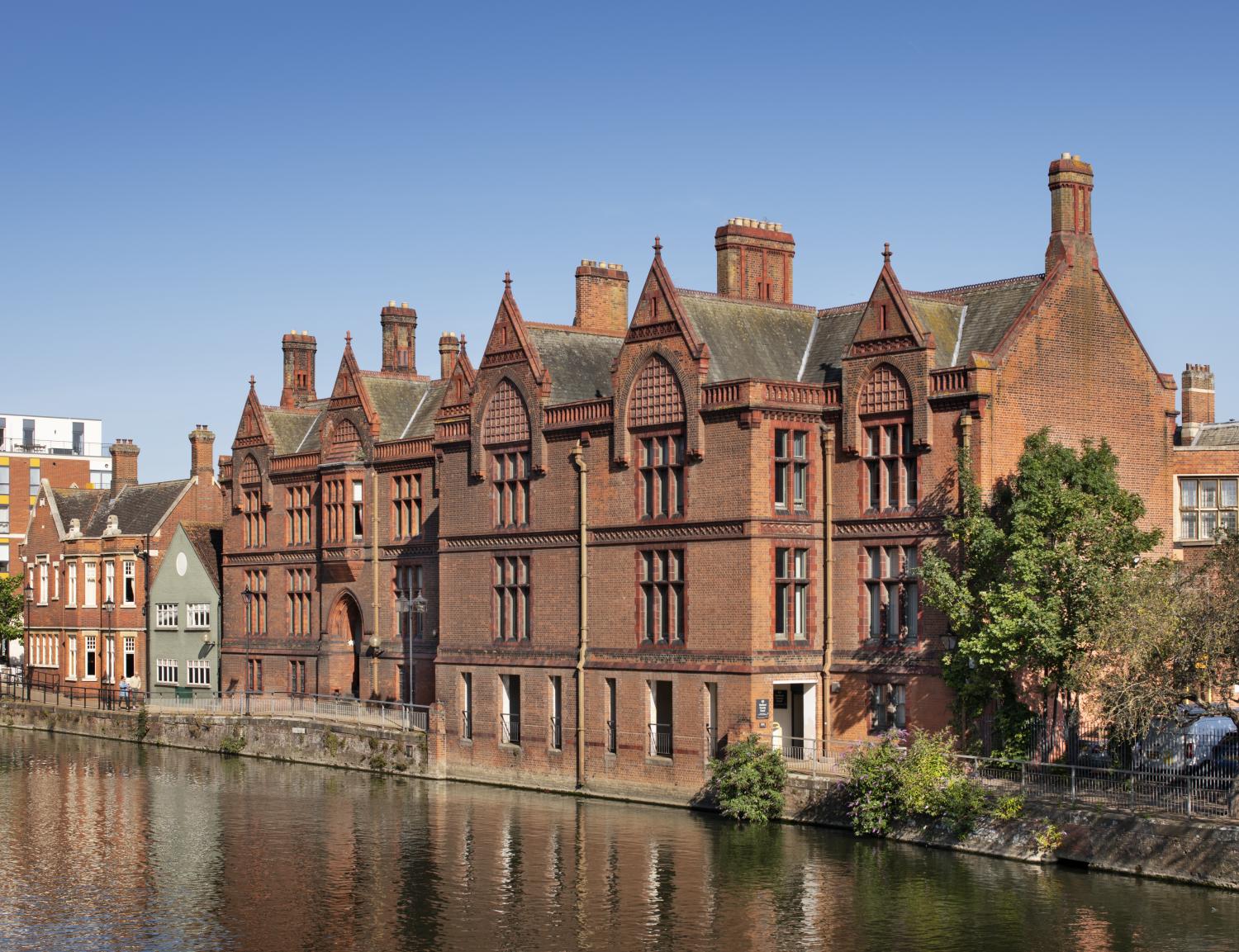 A large red brick building with ornate architectural details, situated on the edge of a river,