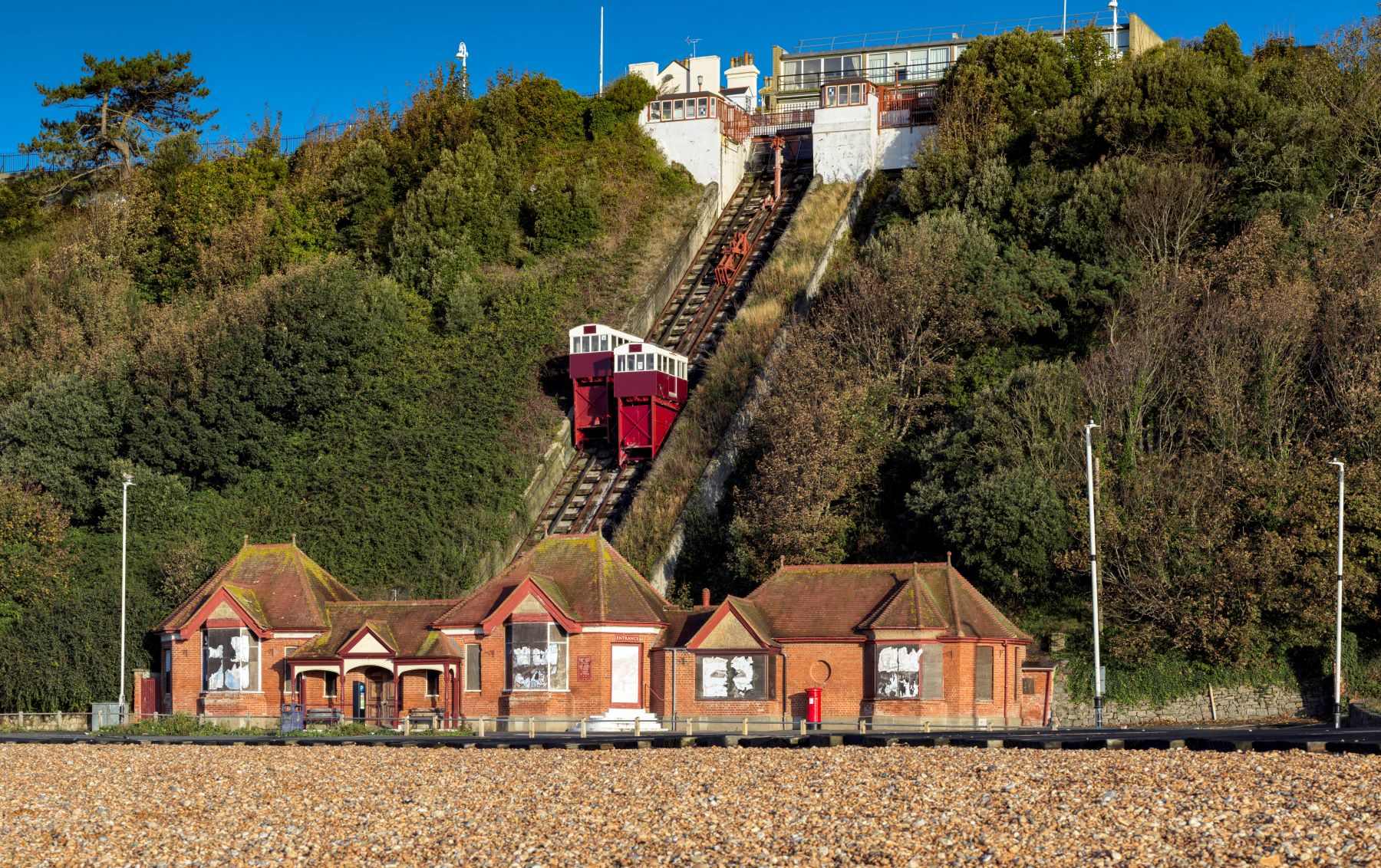 Photo of brick building by a pebble beach with two red lift carts half way up a steep green bank