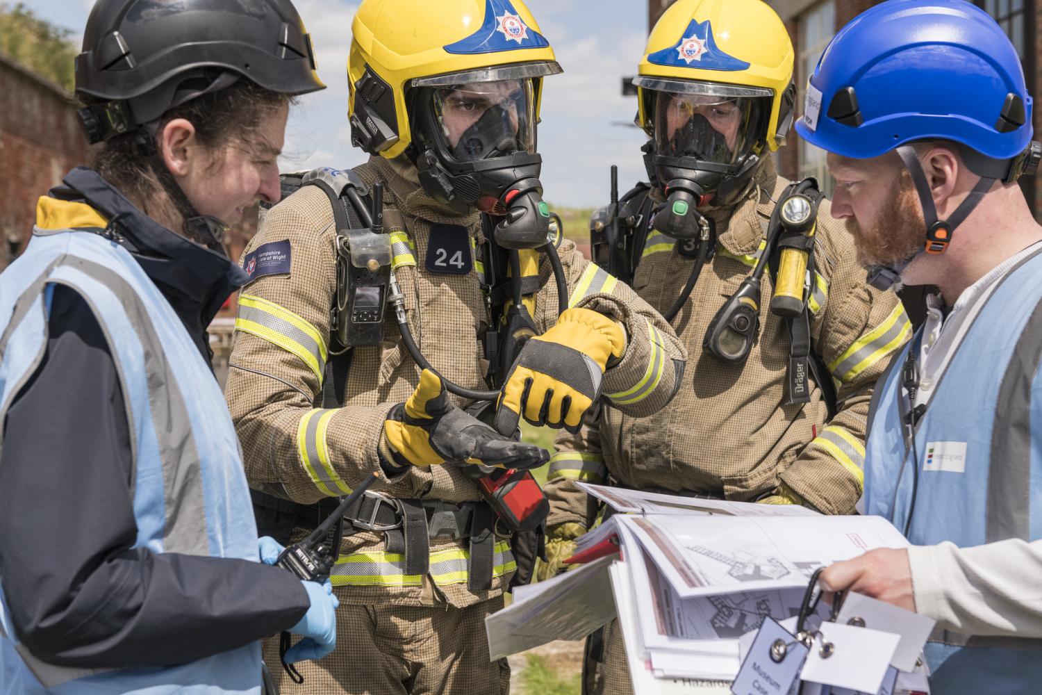 Two firefighters in yellow helmets and wearing breathing apparatus talk to two people in blue high vis and hardhats.