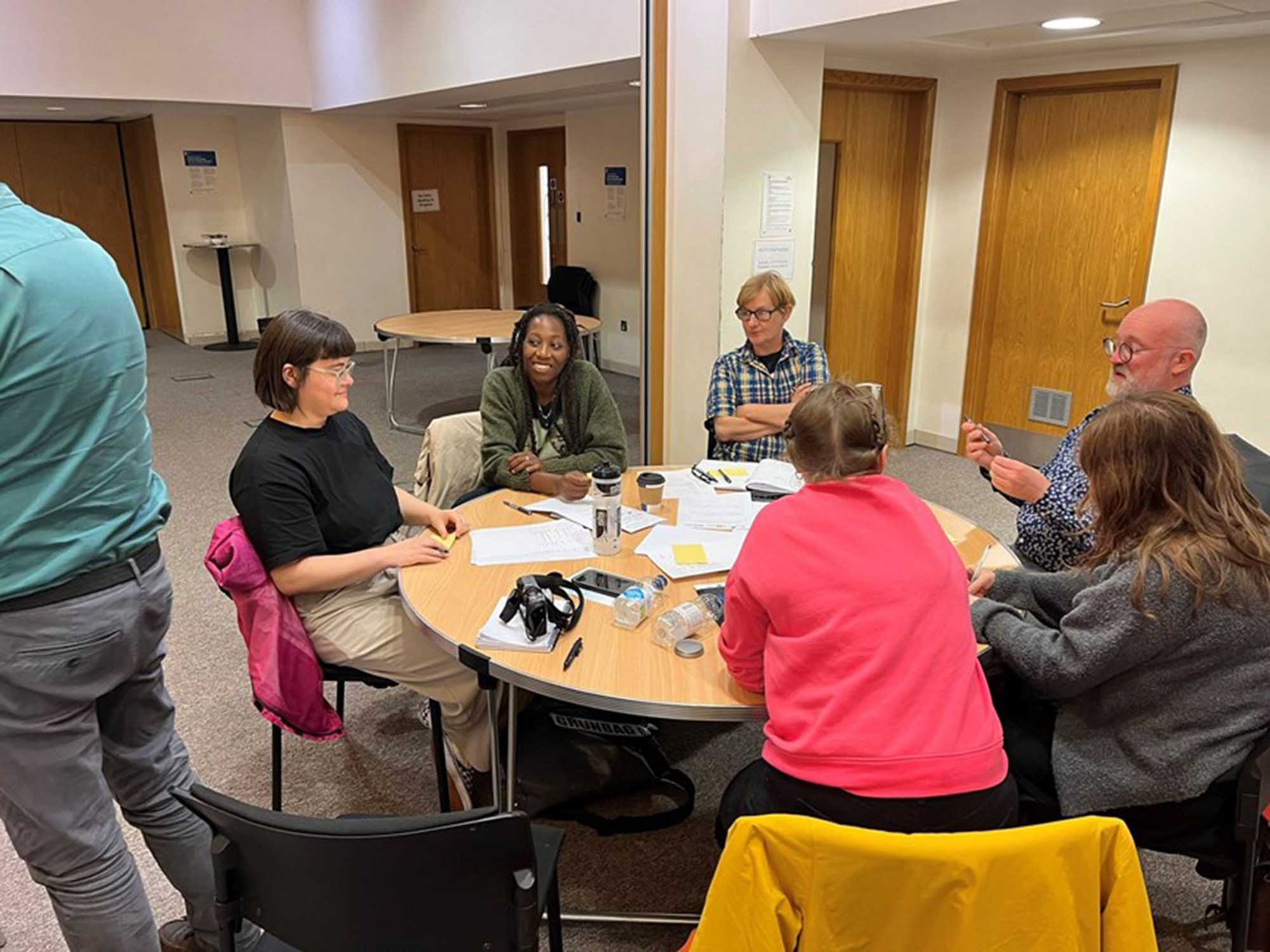 Six people sit around a circular table whihc has scattered papers, pens and bottled water on it. The people are either leaning in, smiling at each other, folding their arms are gesticulating as though they are talking to the group.
