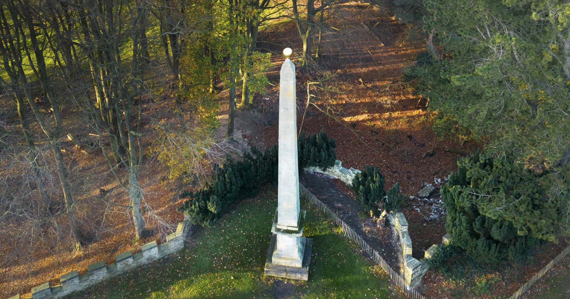 An obelisk monument in a large garden and woodland setting. 