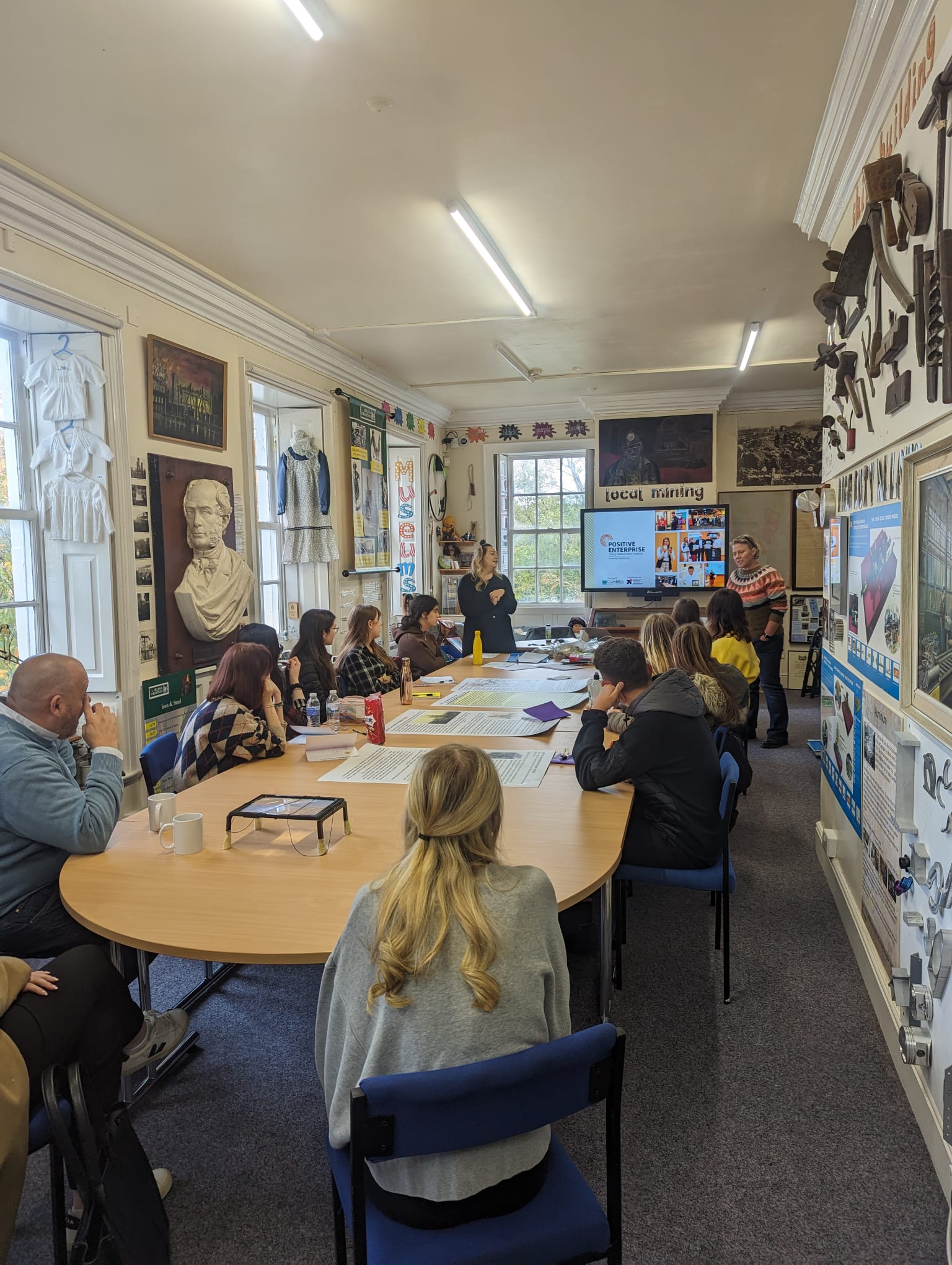 A project group sat around a table observing two speakers giving a presentation.