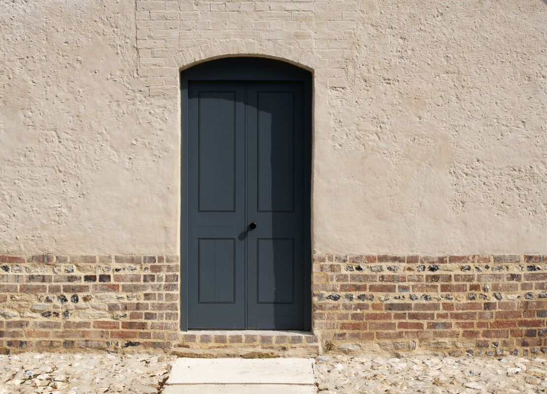 A dark gray door with an arched top is set in a textured beige cob wall with a brick base.