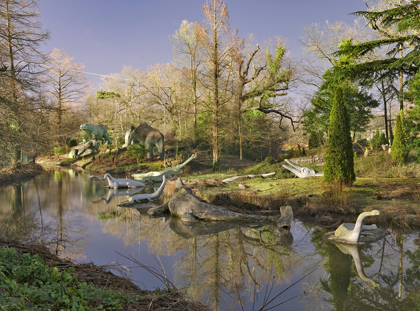 Various concrete dinosaurs around a water feature in parkland. 