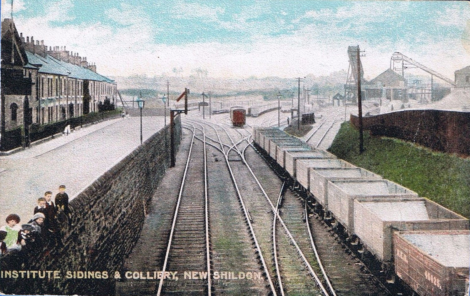 Children looking down from behind a stone wall with houses overlooking the railway with coal wagons and a coal mine in the background