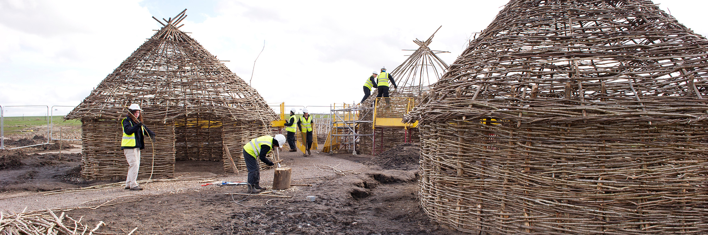 Ground-level image of 6 people in high-visibility clothing and protective helmets building Neolithic-style huts.