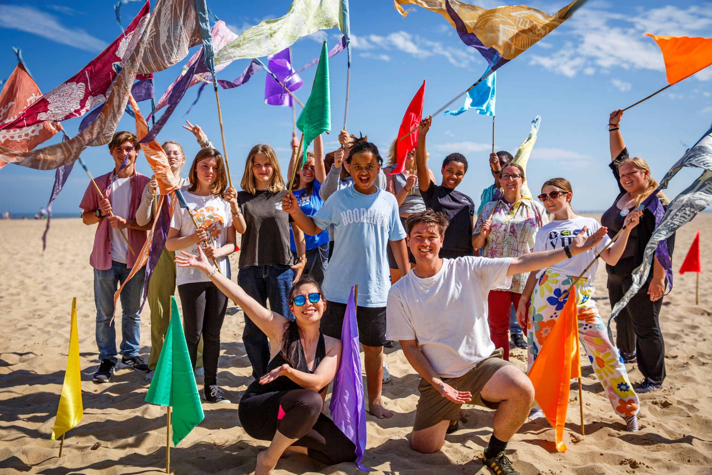 The photo shows a group of young people on a beach with vibrant created flags