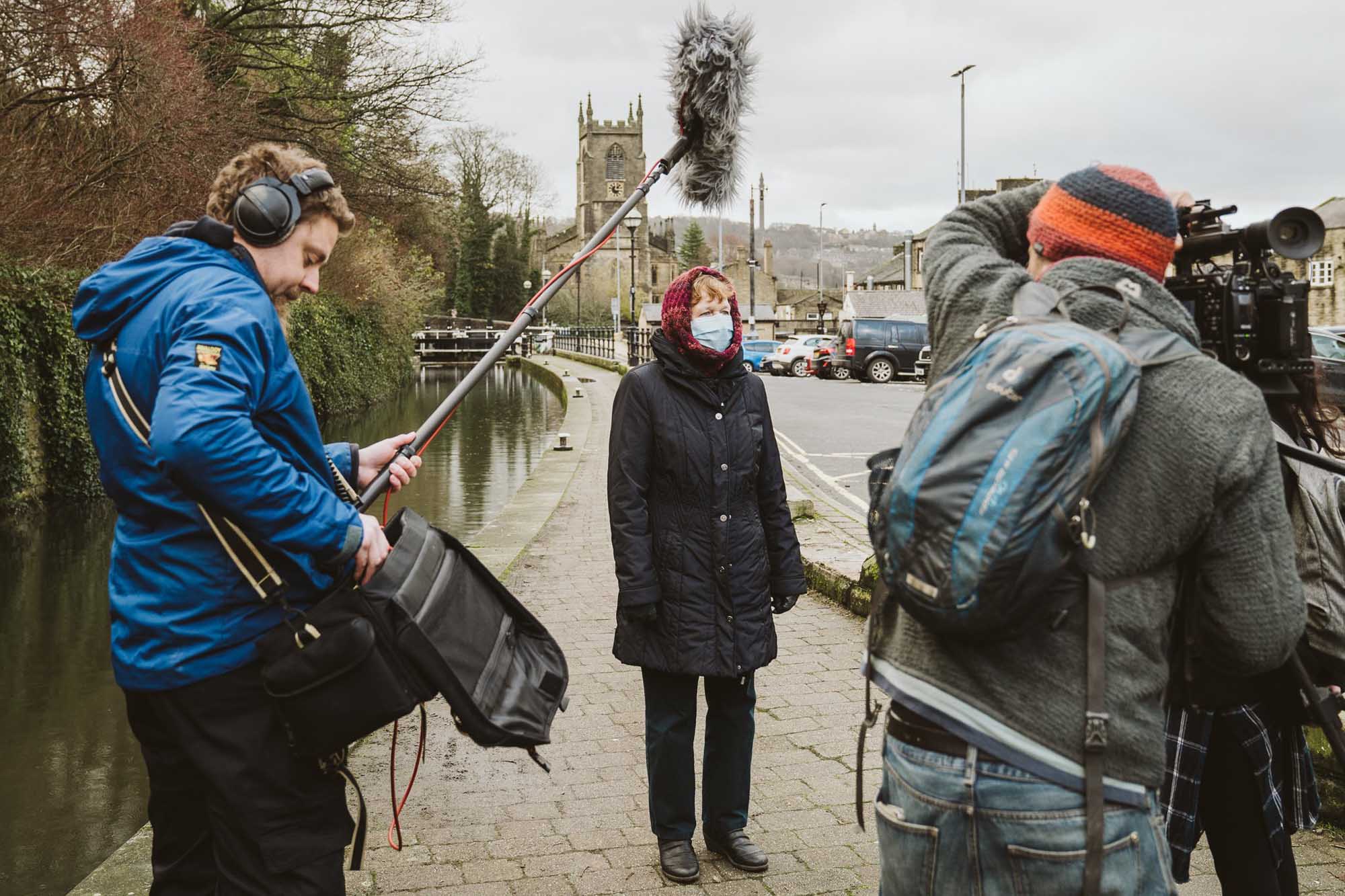 A woman wearing a mask is interviewed with a canal and church in the background. Camera crew are left and right in the foreground.