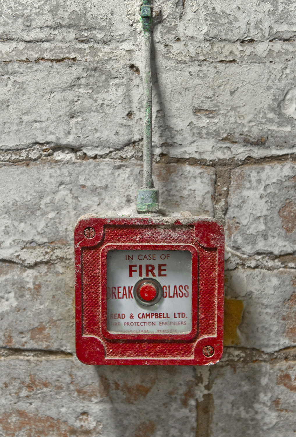 A red metal case surrounds a fire alarm button on a brick wall. 