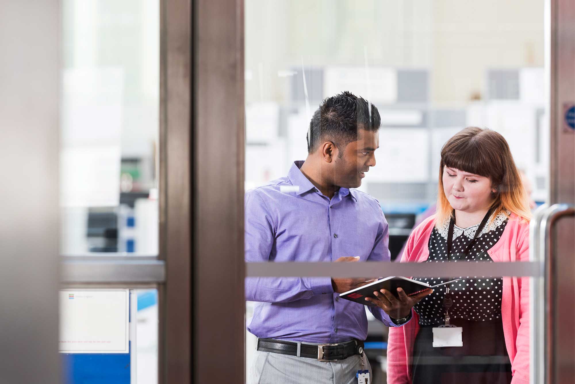 Two colleagues discussing work, viewed through a glass door.