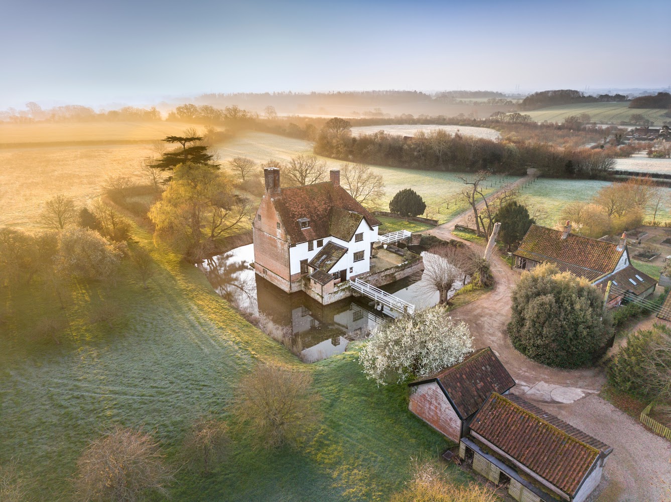An aerial view of Letheringham Lodge shows the building surrounded by a moat in a landscape of fields and trees.