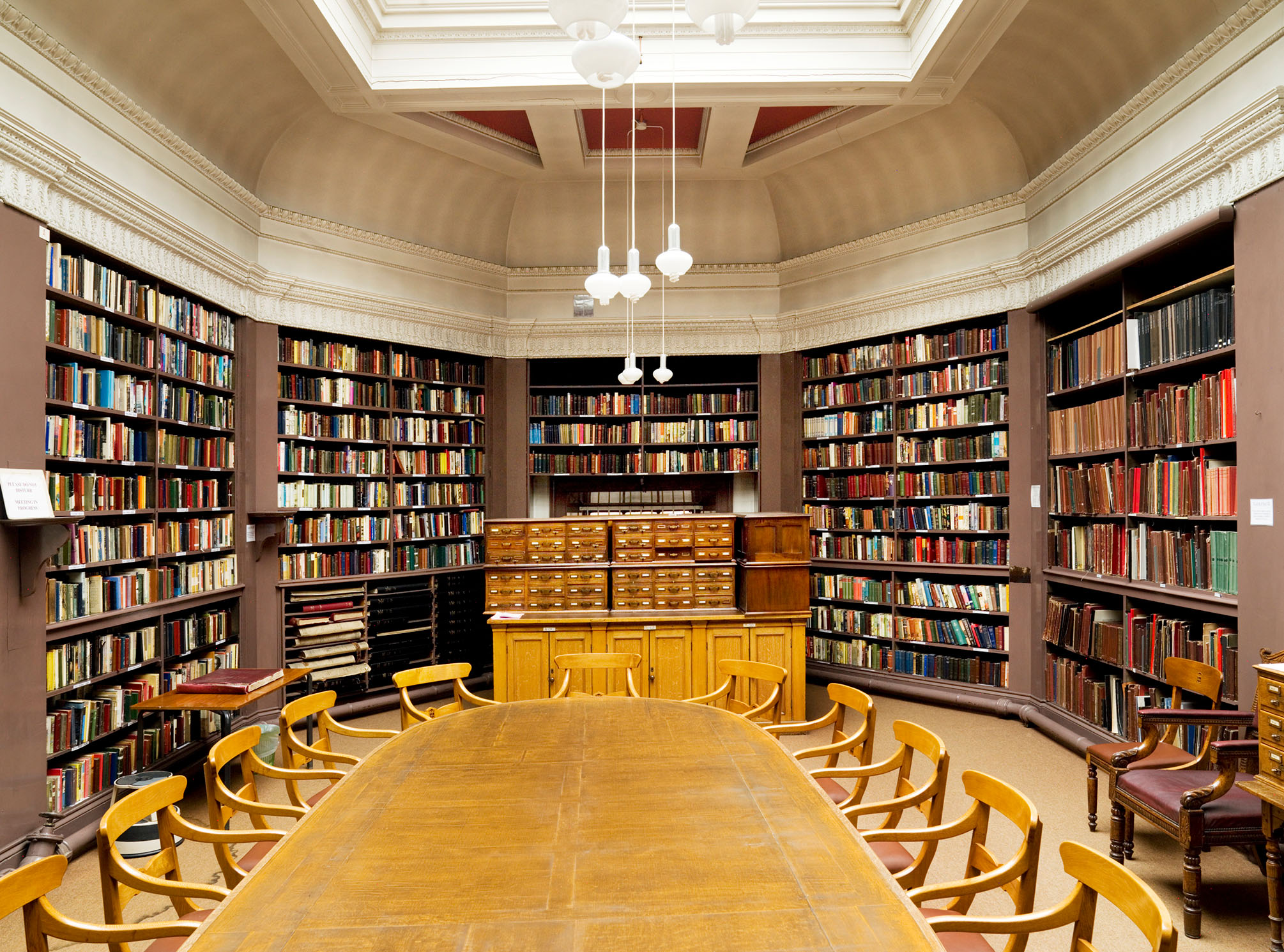 A well-lit library room with wooden shelves filled with books. There is a large wooden table surrounded by chairs in the center, and a set of drawers against the far wall