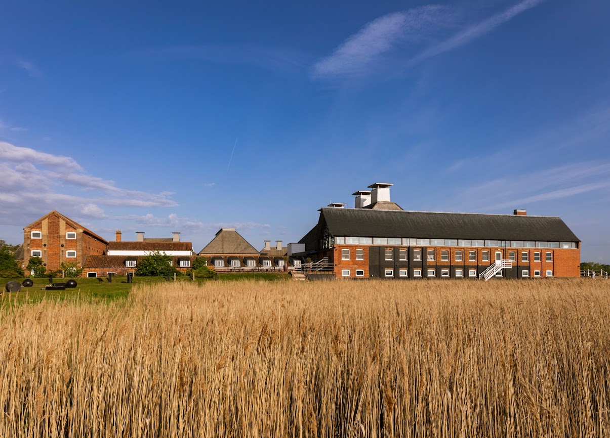 Through wheat fields, in the distance are a collection of red brick buildings, with a large long building to the right, small buildings in the centre and the front of a long building to the left.