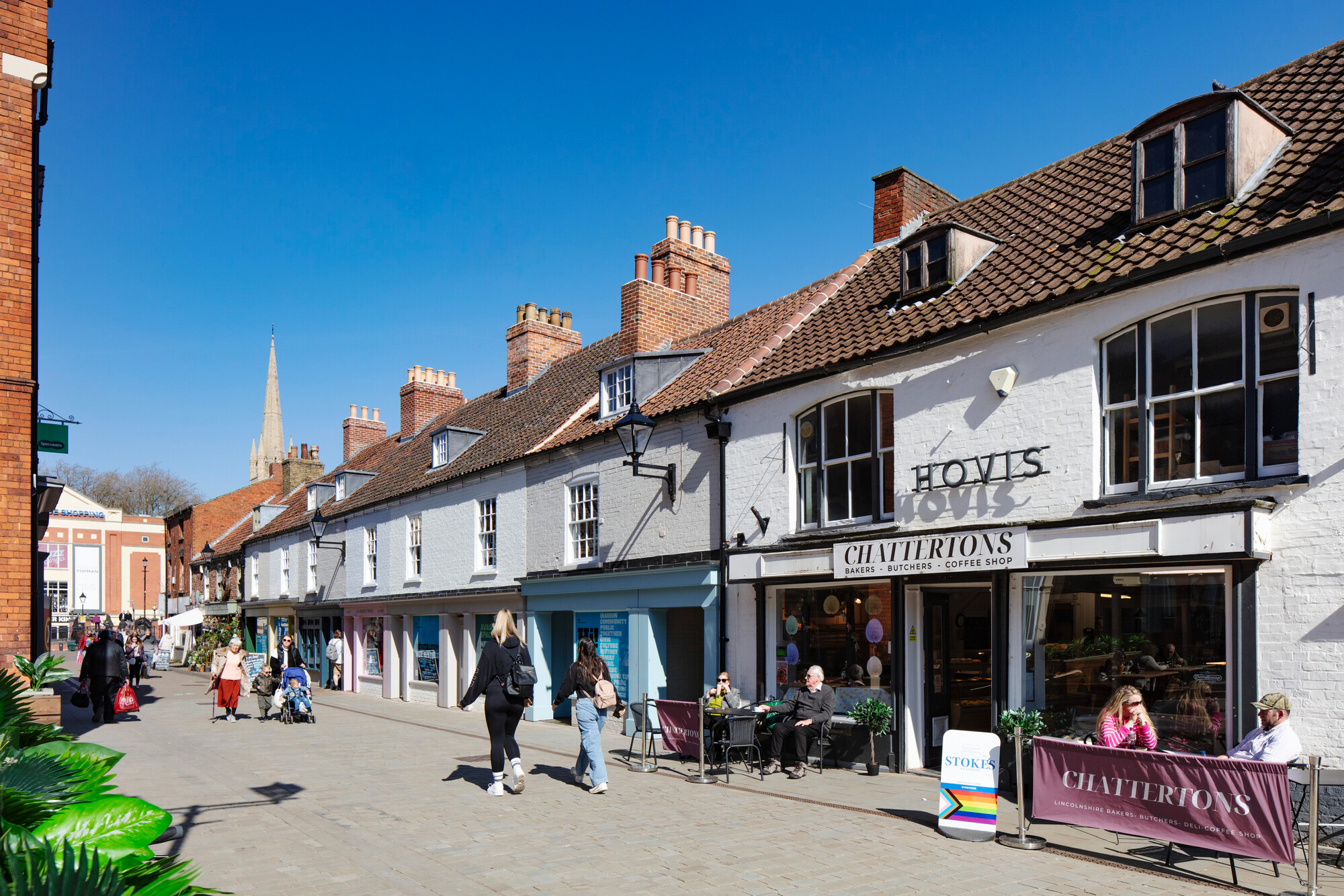 A street scene featuring historic buildings, shops, and outdoor seating. Clear blue skies and a church spire in the background enhance the inviting atmosphere of the area.