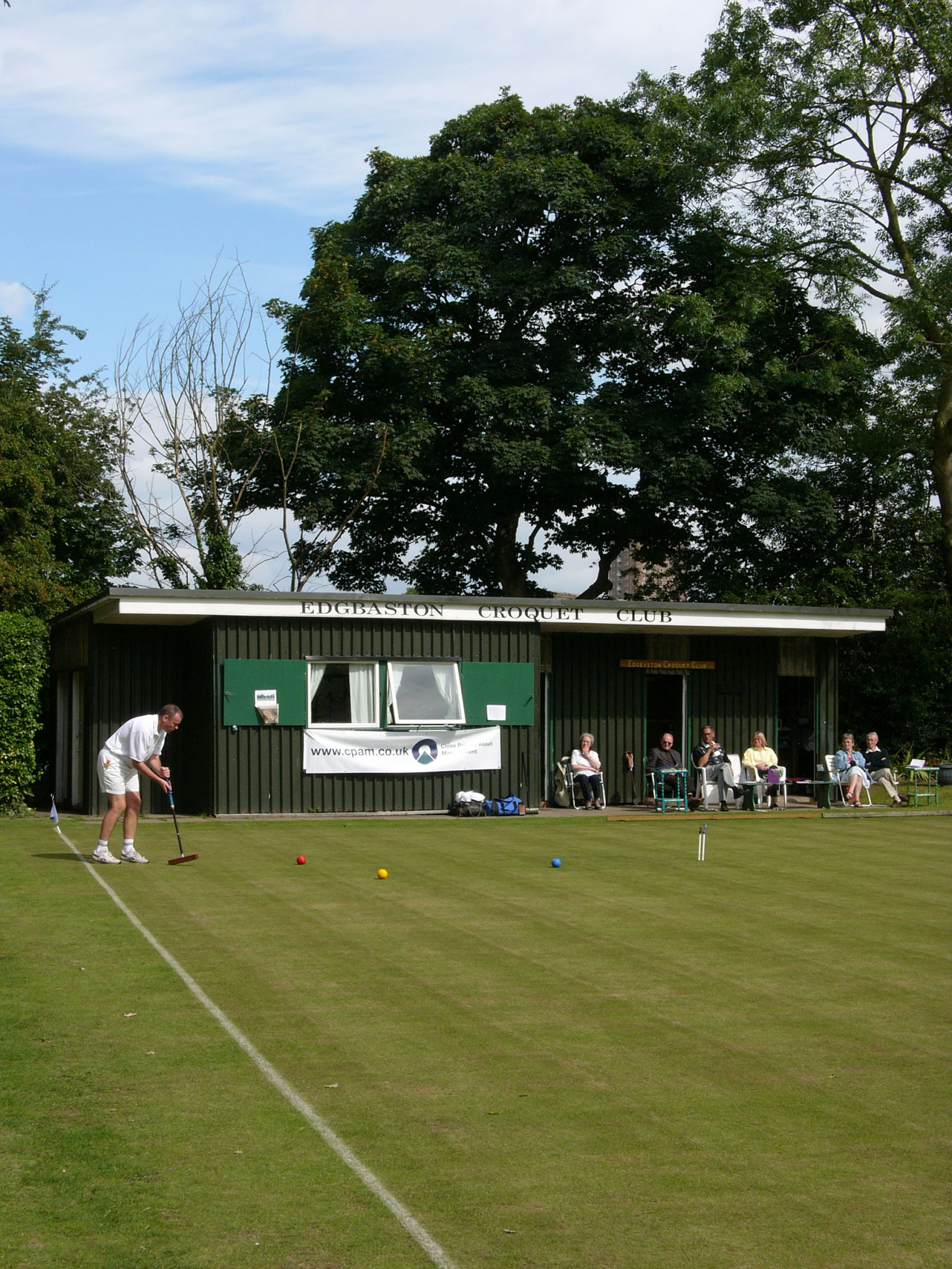 Colour photograph showing a man wearing a white shirt, white shorts and white socks playing croquet. It appears as if he has just hit a red ball towards a hoop, and close to a yellow ball and a red ball. In the background is a small, single-storey pavilion. Along the top of the pavilion are painted words reading: 'Edgbaston Croquet Club'. Six spectators sit on chairs in front of the right side of the pavilion. Behind the pavilion are a variety of trees.