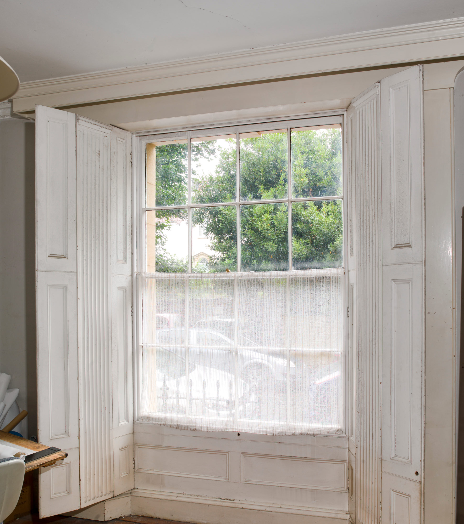 Sash window with view onto parked cars through a net curtain. Trees are visible through the plain glass top half of the window. On either side there are wooden shutters.
