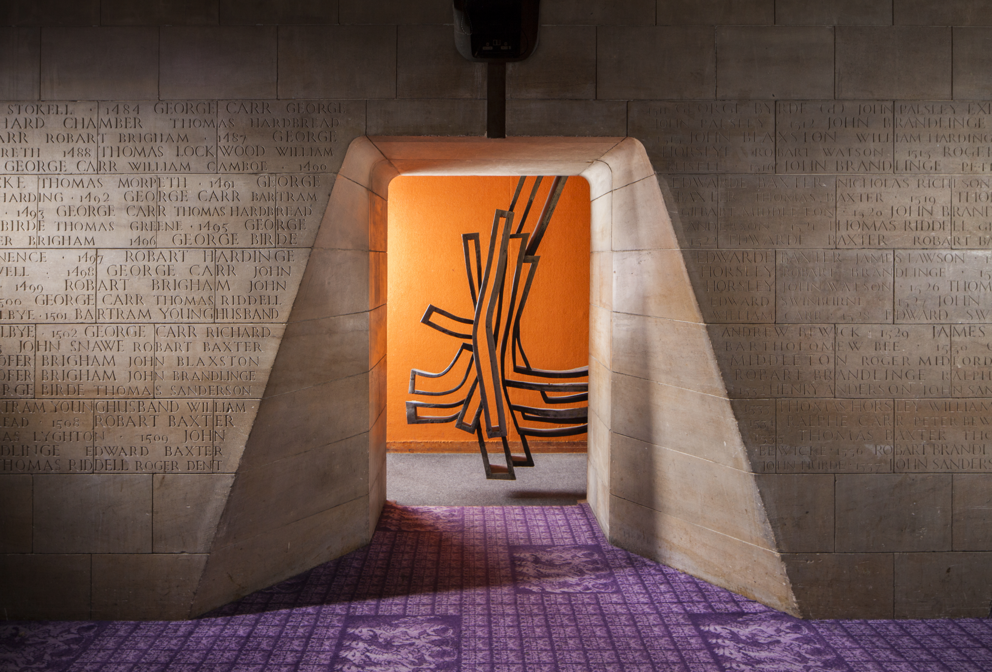 Stone walls covered in engraved names on a purple carpet with an archway leading to a bright orange room.
