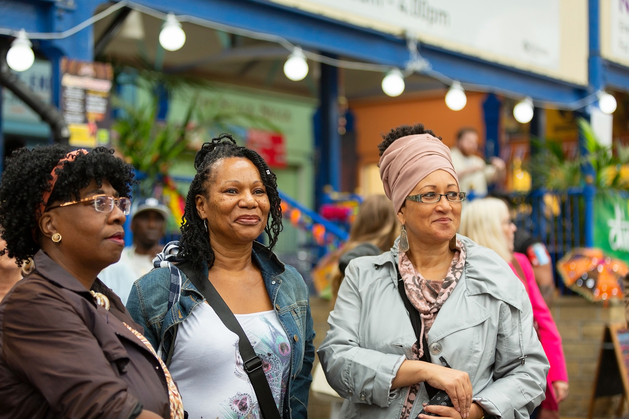 Three women smiling and looking to the side at an indoor event with colourful decorations in the background.