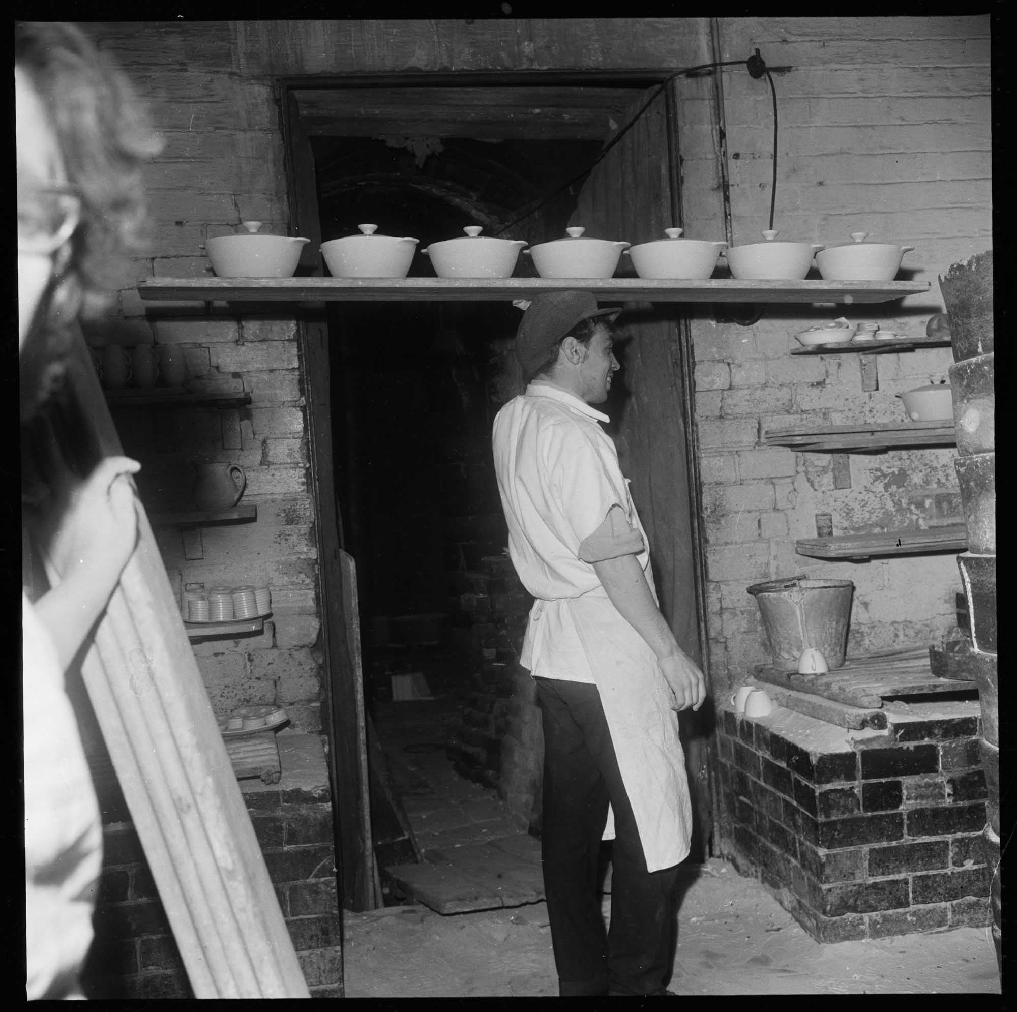 Man wearing cap and apron carries seven unglazed pots with lids on a plank on his head.