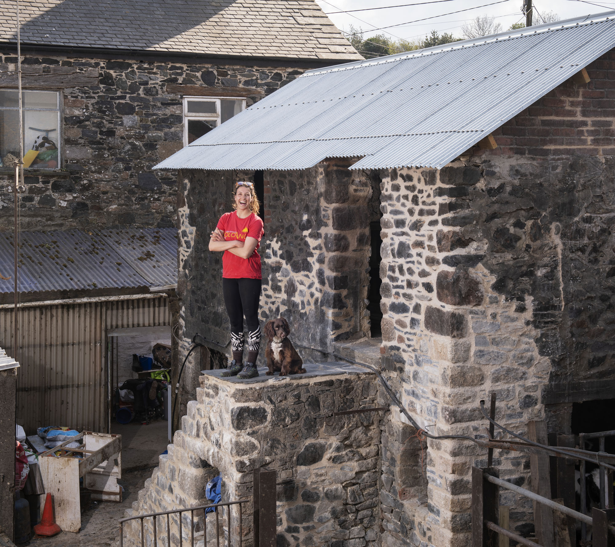 Colour photo of a woman standing at the top of stone steps, accompanied by a dog. The setting features a mix of rustic buildings and construction materials.