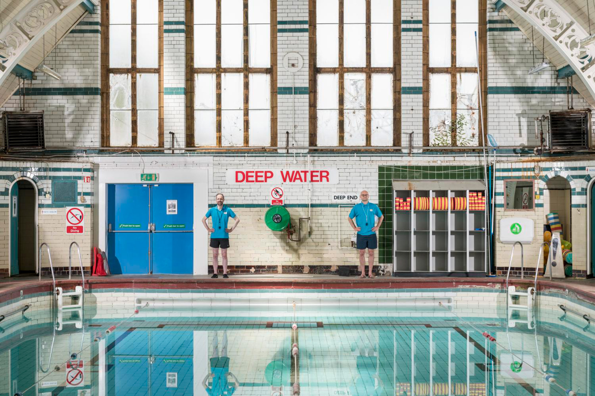 Two lifeguards stand beside an indoor pool marked "deep water," surrounded by tiled walls and large windows.