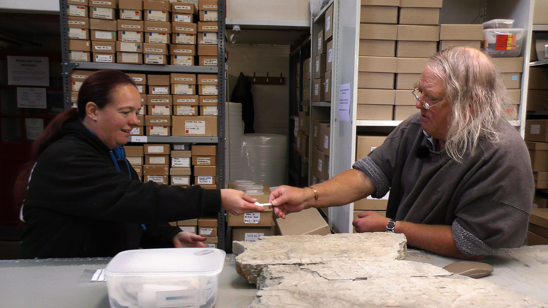A woman and a man discussing archive objects set out on a desk, in the background are boxes of further archives on shelves.