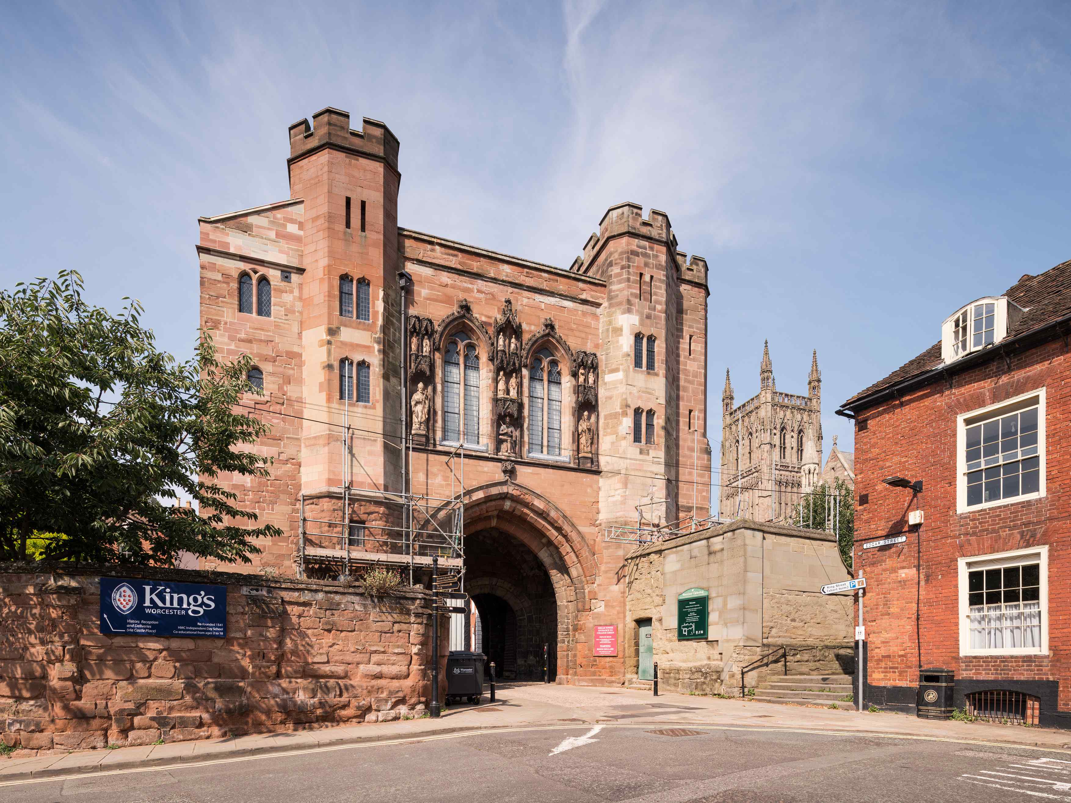 Red sandstone gate tower with large arched gateway on ground level. Church tower is visible on the right  in the distance in between the tower and a red brick house on far right.