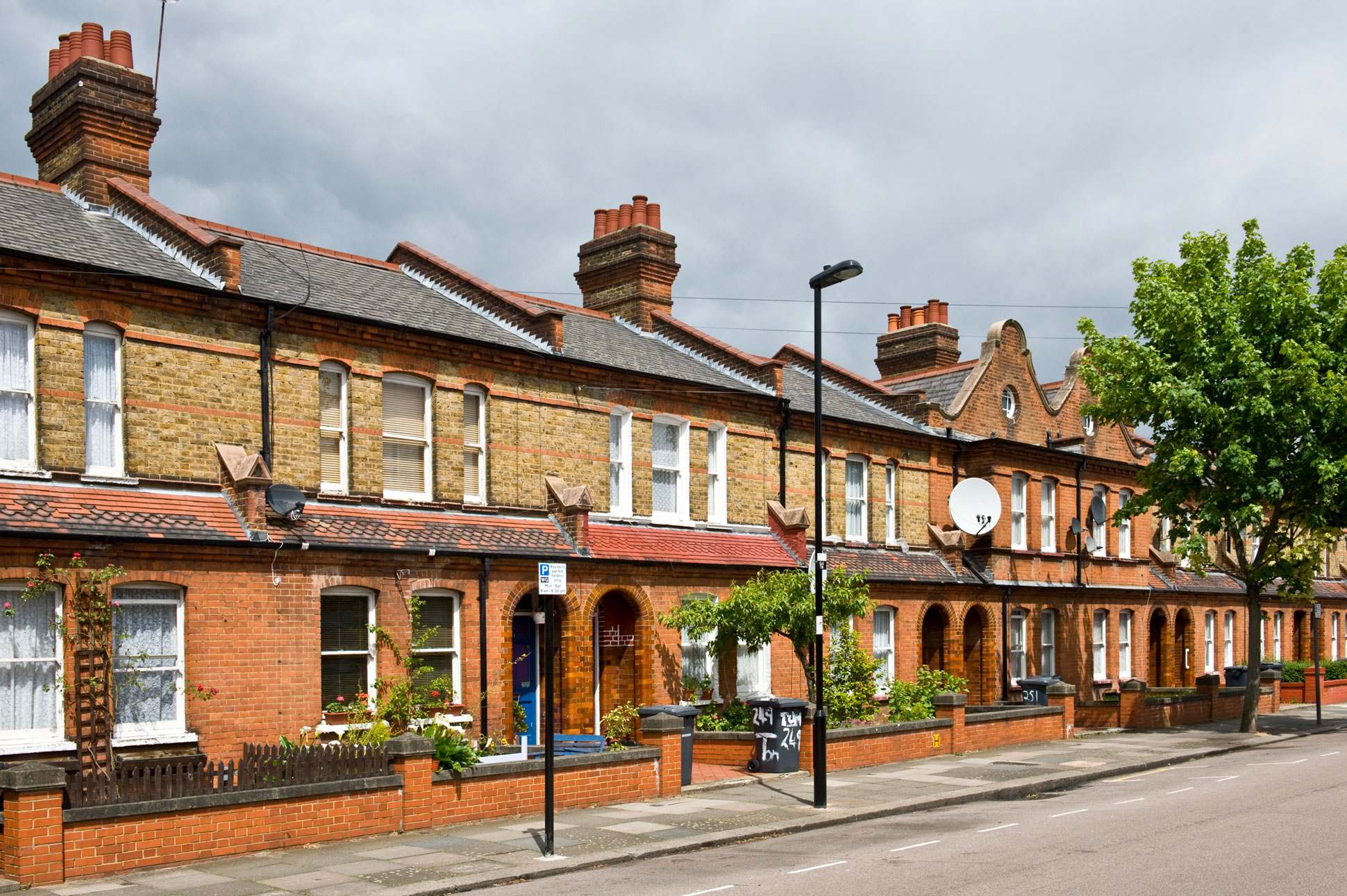 A row of red- and yellow-brick two-storey terraced houses, some with wheelie bins visible over the low wall bordering their front gardens.