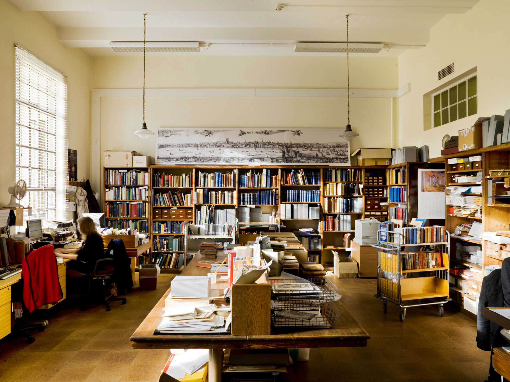 Office with book shelves, a book trolley and central table piled with paperwork and more books. A woman sits at a desk under a window on the left hand wall.