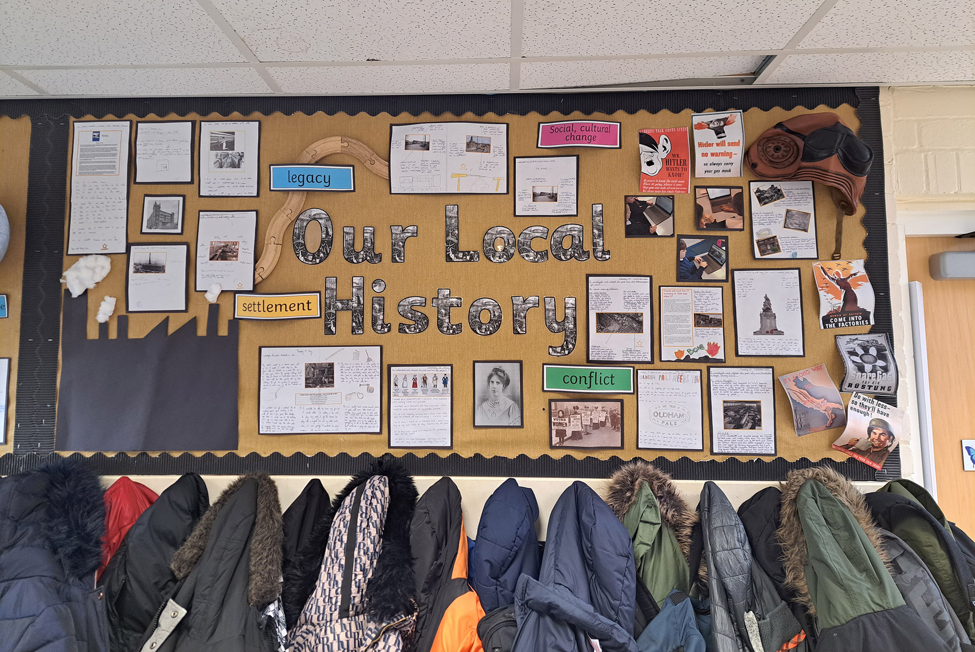 Photograph of a school display board titled "Our Local History" displaying various documents, photos, and colourful labels such as "legacy," "settlement," "conflict," and "social culture change." The board highlights key historical themes and events with organized sections and visual elements, situated above a row of hanging coats.