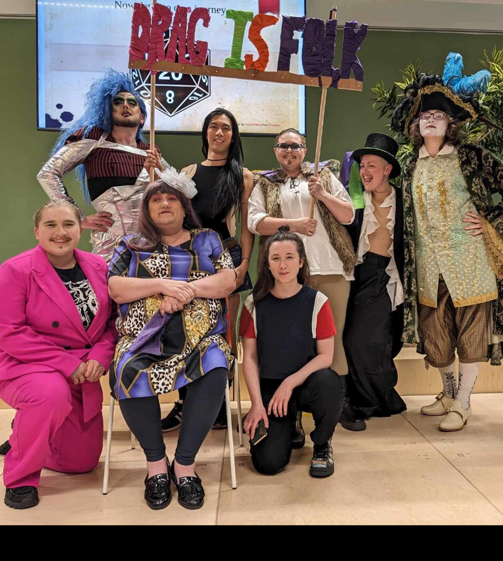 8 people in costume hold a handmade sign that reads 'Drag is Folk'