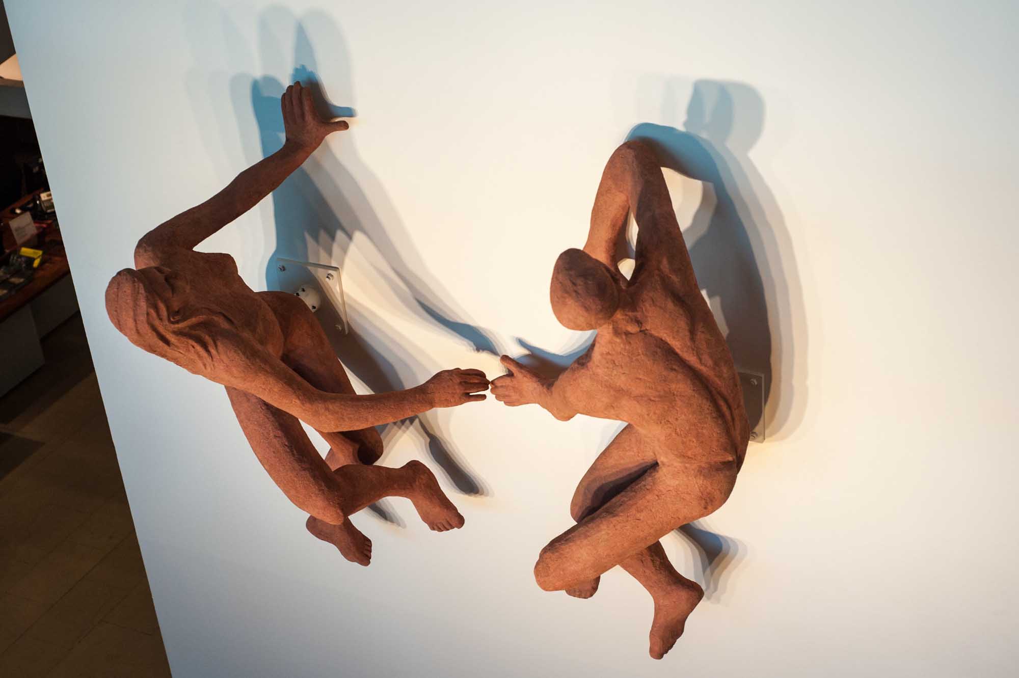 Close-up photo of two red figures reaching towards each other. Their shadows are cast on a white background.