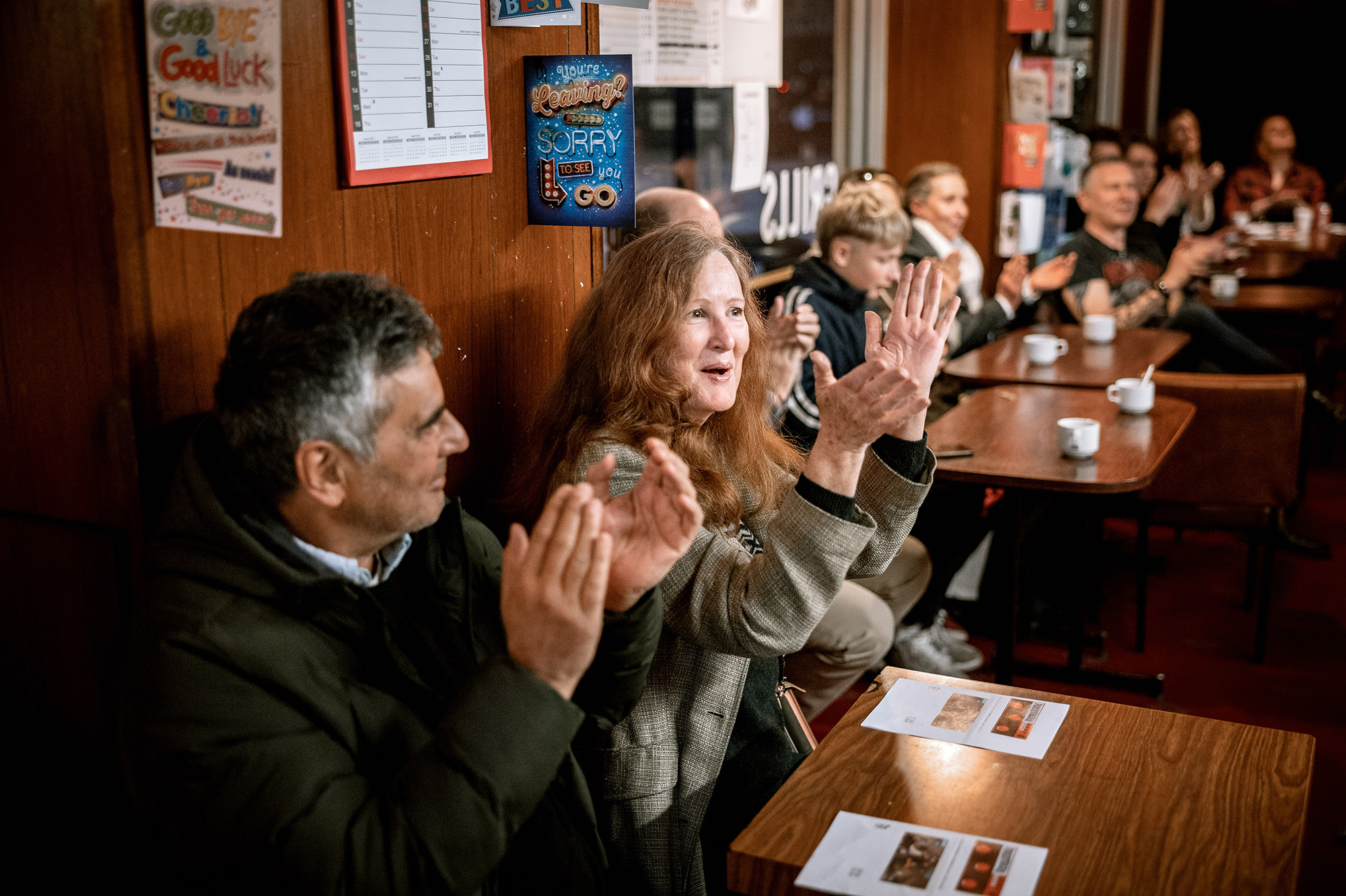 Eleven people of various ages sit at a number of small wooden tables repositioned to be in a row in a cafe or a pub. The people are facing in one direction, looking towards something out of shot. There are cups of coffee on the tables and the people are clapping.