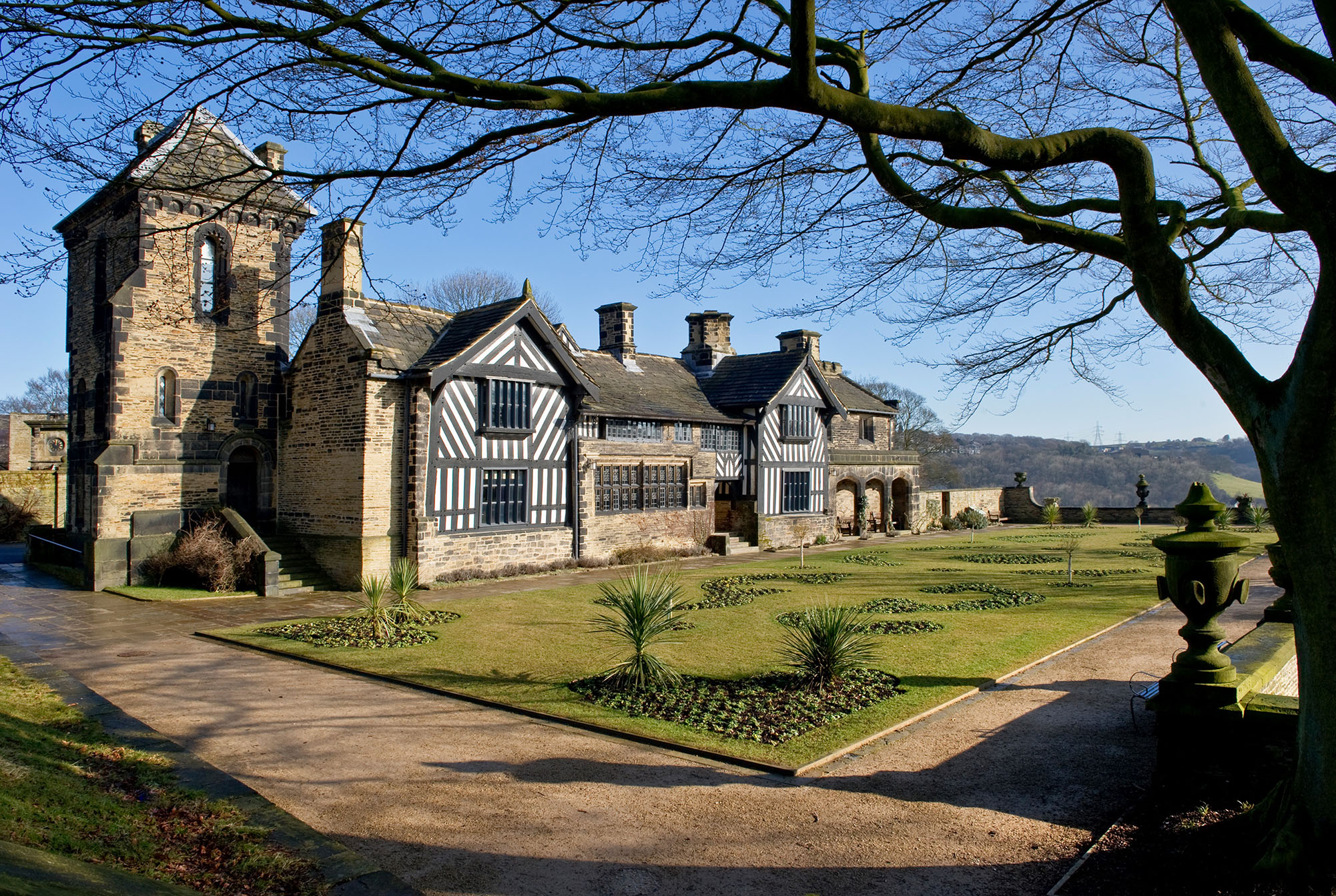 Photo of Shibden Hall, a long house with a mix of timber-framed and stonework walls. A stone tower stands slightly behind the house, closest to the camera. There are manicured gardens before the house and the path is lined with stone urns.
