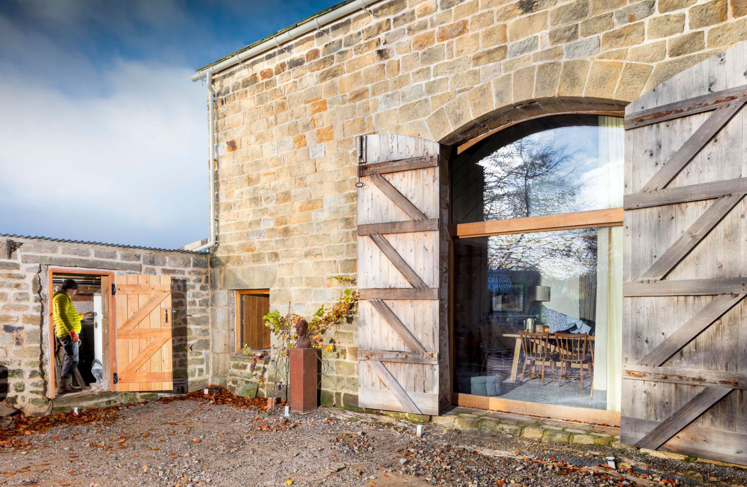 Detail of the exterior of a converted stone barn with a large window. Adjacent to this is a one-storey stone outhouse in which a person stands next to a heat pump.