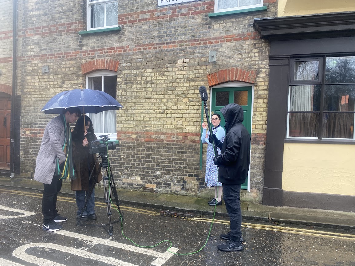 Against a historic terraced house, a young film crew stand in the street.  A woman in  vintage dress stands in the background, smoking a cigarette.