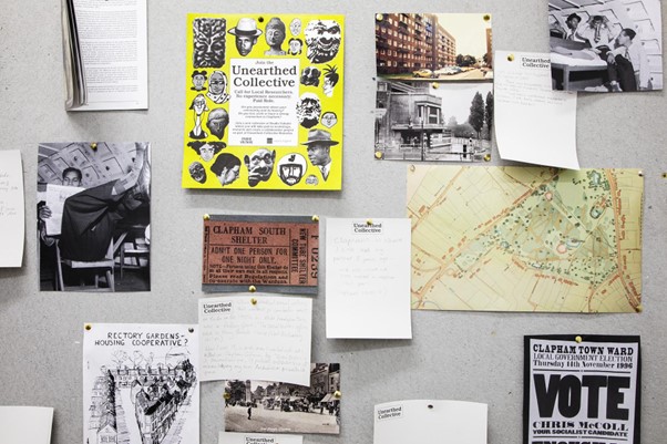 A bulletin board displaying various black and white photographs, a map, handwritten notes, and a yellow booklet titled "Unearthed Collective".