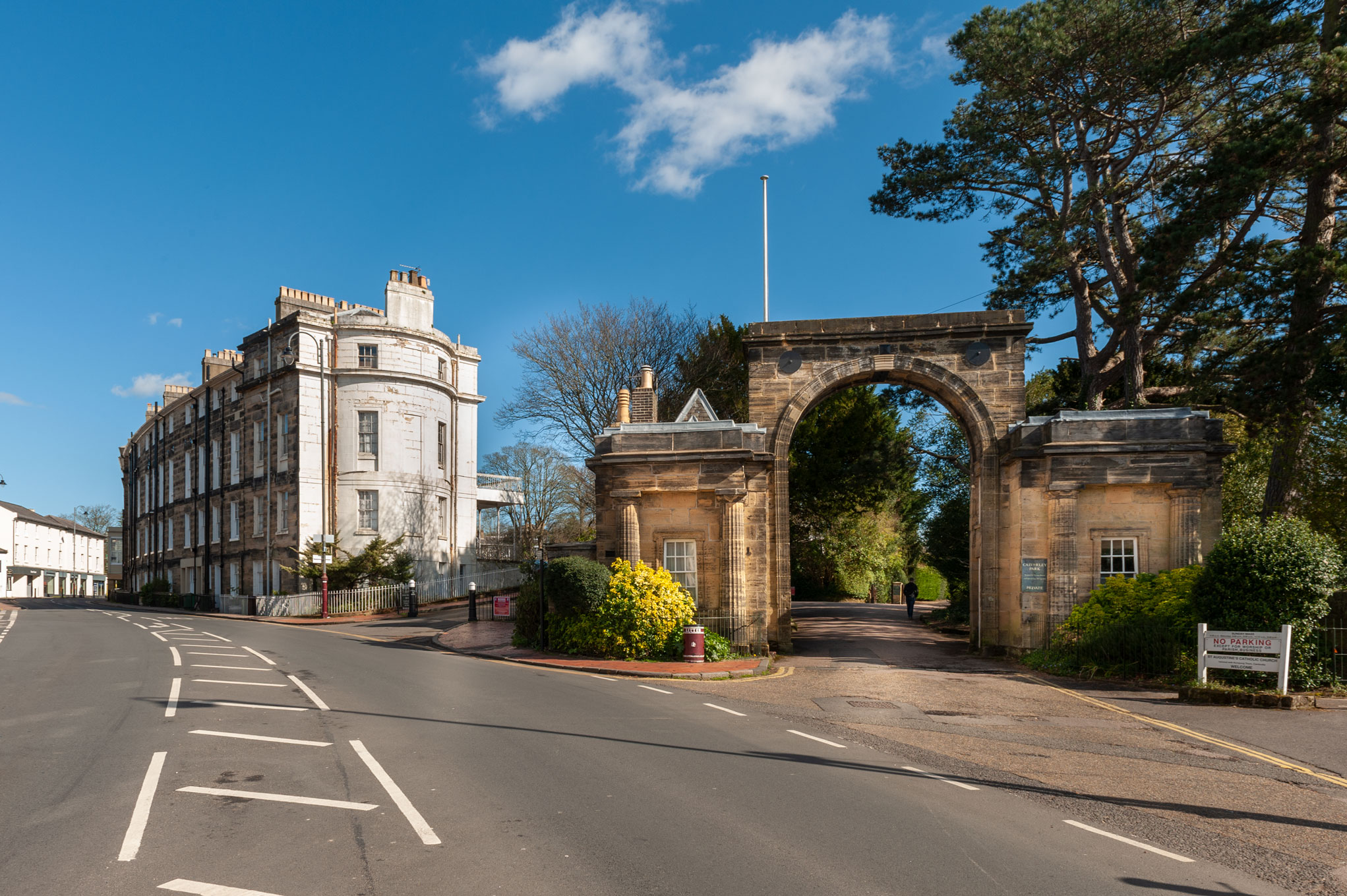 Colour photograph of a stone gateway with trees beyond and a curved block of white-coloured buildings to one side.  

