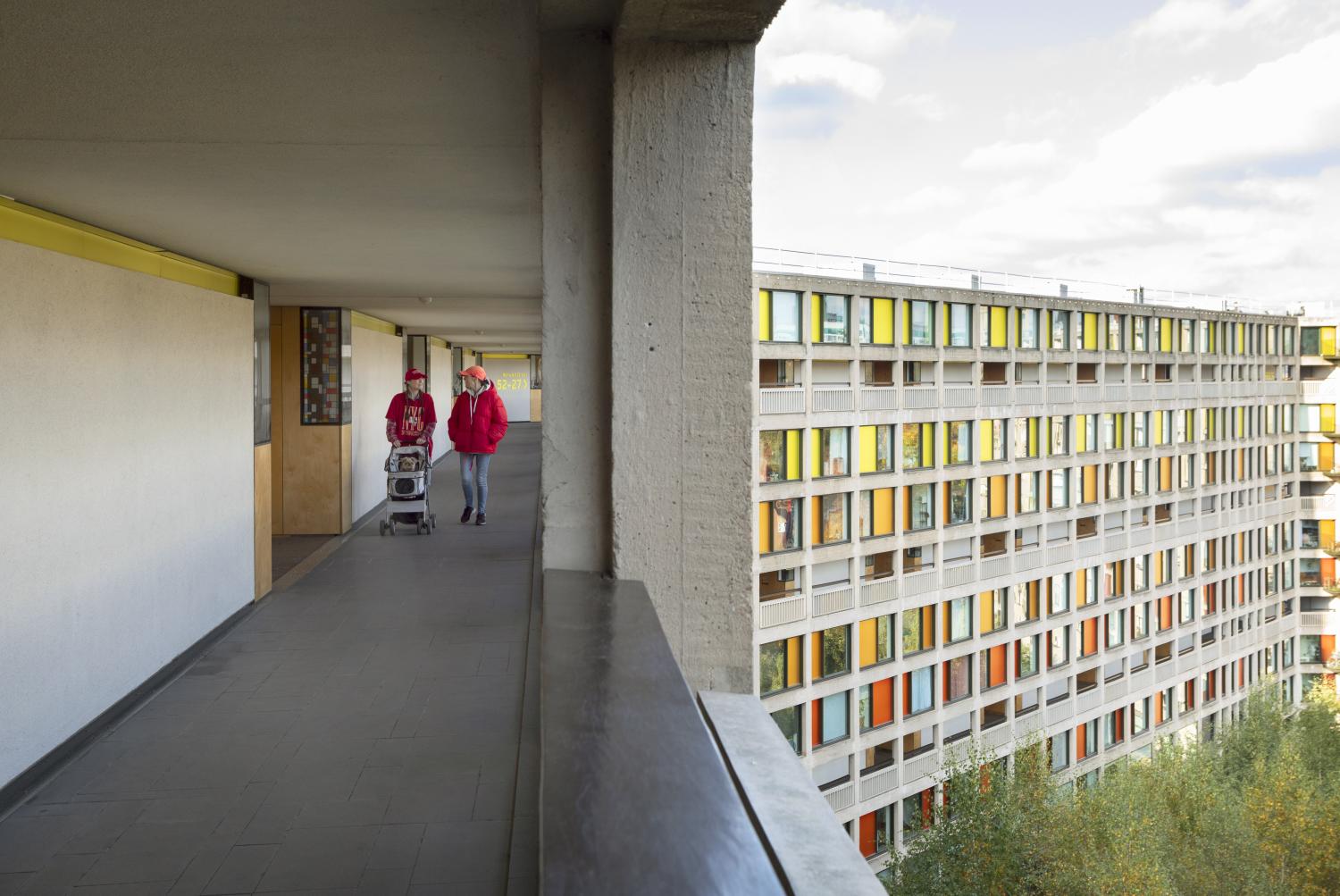 Two people in red jackets walk down a corridor, pushing a small cart. They are on an upper floor of a building with a large windowed facade visible in the background. The sky is partly cloudy, and lush greenery surrounds the area below.