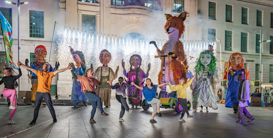 A line of people dancing in front of a line of street carnival puppets, with pyrotechnics in the background.