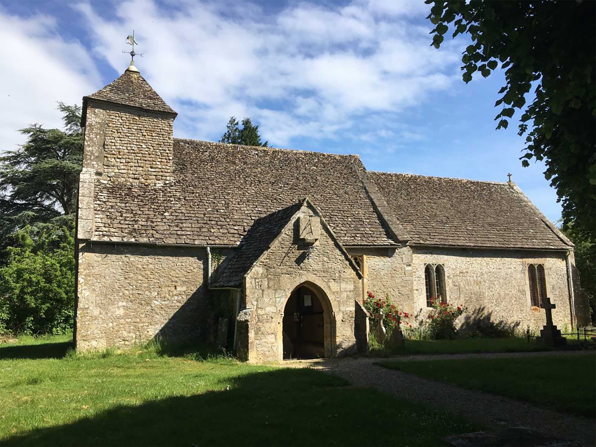 Landscape photograph of the front exterior of St Michael Harnhill. It is a grey stone church with green grass and a blue sky.