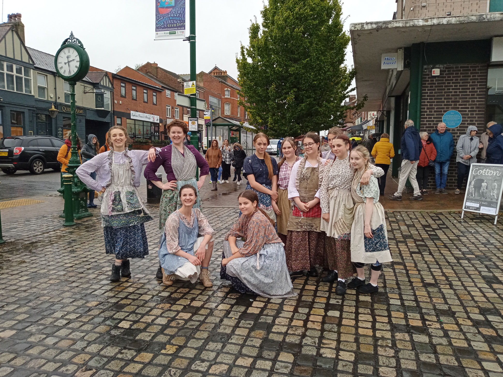 Young women and schoolgirls dressed as Victorian or Edwardian mill girls pose together for a photograph in a pedestrian precinct.