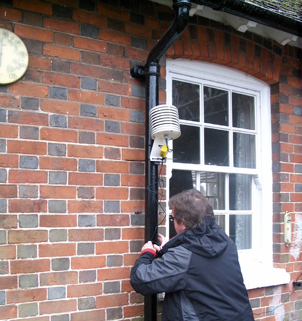 A man attaches a monitor to a drainpipe on the outside of a red brick building.