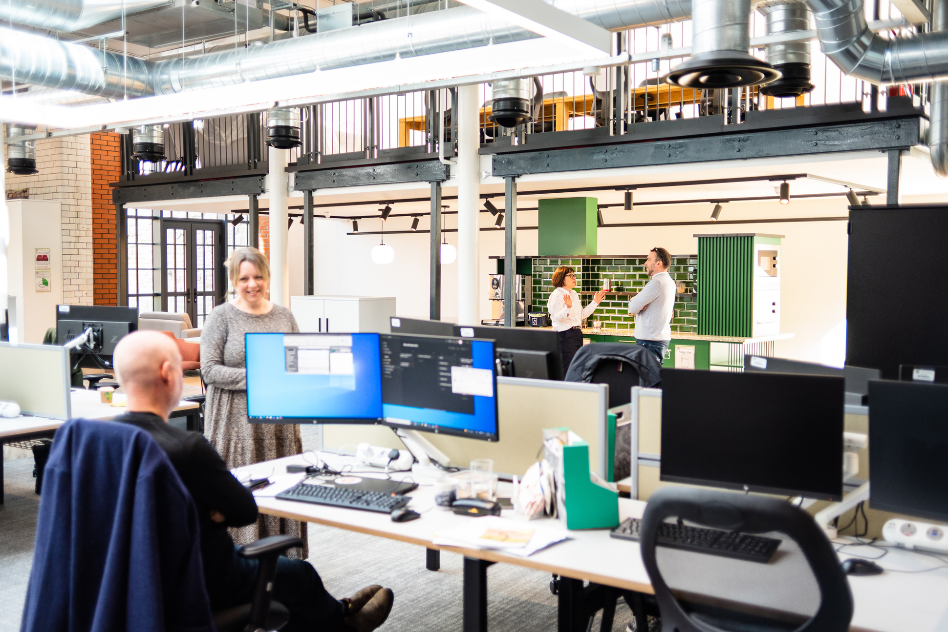 Open plan office showing workers in conversation between desks and in a kitchen area in the background.
