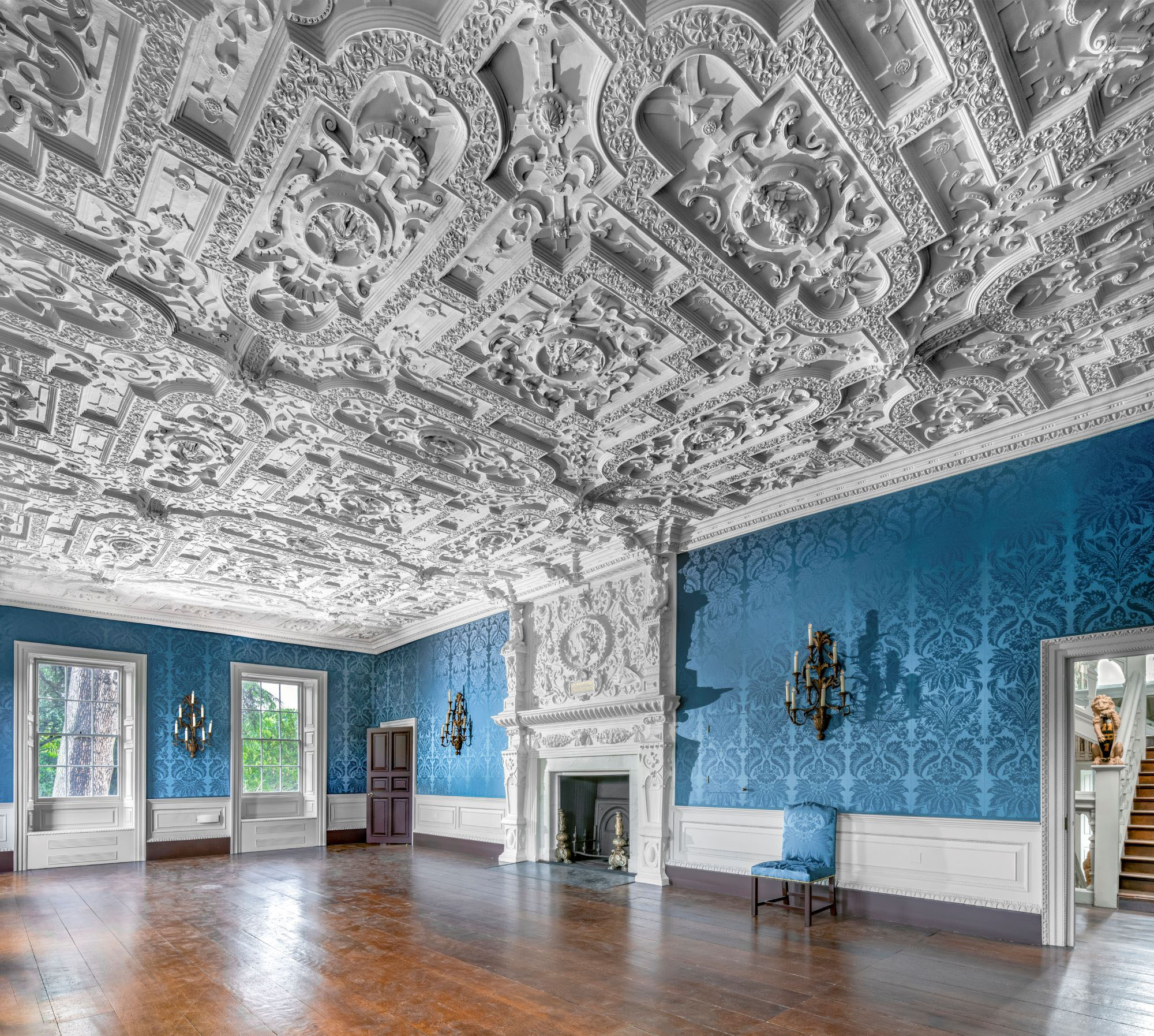 View of a room with a decorative white plaster ceiling, blue walls and a wooden floor. There is an ornate fireplace in the centre of one wall.